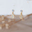 Gold teardrop charm stud earrings with pavé crystals, displayed on a soft neutral fabric with dried floral accents in the background.