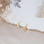 Elegant gold teardrop charm stud earrings with pavé-set cubic zirconia, displayed on soft cream fabric with dried floral accents.