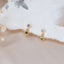 Elegant gold drop earrings featuring a round green gemstone charm paired with a clear cubic zirconia stud, displayed on cream textured fabric and soft satin background with dried floral accents.