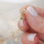 Close-up of a hand holding a dainty gold stud earring with a green gemstone charm and clear crystal top, shown with glossy pink-speckled nails and a soft neutral background.