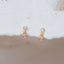Gold drop earrings with round pink gemstone charms and sparkling clear studs, displayed against soft white fabric background.