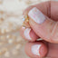 Gold stud earring with a dangling pink gemstone charm and clear crystal top, shown between fingers to highlight size and detail.