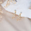 Gold snake stud earrings with wavy crystal-encrusted charms and green gemstone eyes, displayed on soft fabric with dried floral decor.
