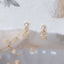 Small gold star charm stud earrings with clear cubic zirconia, photographed against a soft fabric background with dried floral accents.