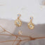 Gold sun charm stud earrings with clear crystal tops and eye detail in the center, displayed hanging on a textured white surface with delicate dried flowers in the background.