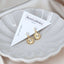 Pair of gold sun charm stud earrings with clear crystal tops and eye detail, displayed on BenittaMoko Jewelry card in a scalloped ceramic dish