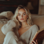 Blonde woman with natural makeup and soft waves, wearing gold hoop earrings and a cozy beige knit sweater, sitting on a couch and holding a book in a warm, relaxed setting.