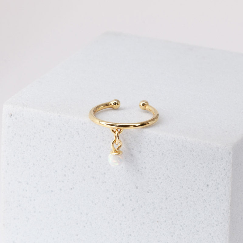 A gold-colored ear cuff earring with a white opal ear cuff, displayed on a white cube-shaped display. The ear cuff is designed to fit comfortably around the ear without requiring a piercing and features a beautiful white opal stone that adds a touch of elegance and sophistication to the accessory