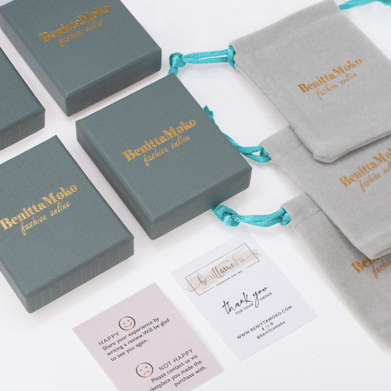 Packaging from BenitaMoko jewelry, featuring gray gift boxes and bags with the company's gold-stamped logo. The packaging provides an elegant and sophisticated look to the brand's jewelry products and is suitable for gifting or personal use