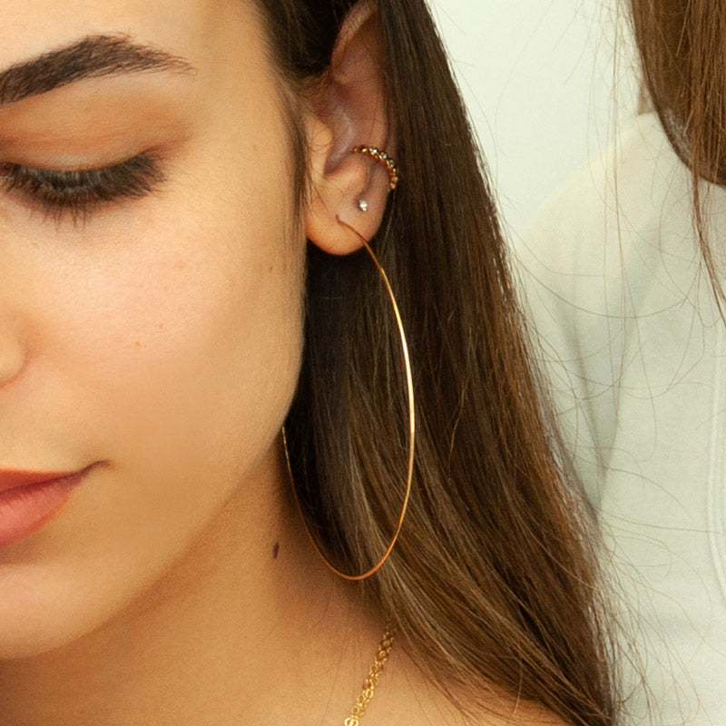 A woman with brown hair wearing gold hoop earrings with a diameter of 3 inches. The earrings are designed in a classic circular shape and feature a shiny, reflective surface. They are worn through pierced ears and add a stylish accessory to the woman's outfit