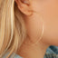 Woman wearing gold hoop earrings with a diameter of 3 inches. The earrings are designed in a classic circular shape and feature a shiny, reflective surface. They are worn through pierced ears and add a stylish accessory to the woman's outfit