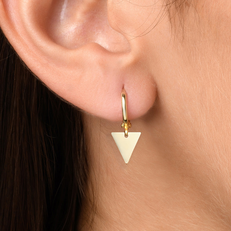 A woman with brown hair wearing gold hoop earrings with a geometric triangle charm. The hoop earrings feature a sleek, minimalist design with a delicate triangle charm that adds a touch of sophistication and style to the woman's look. The gold plating gives the earrings a luxurious and elegant look that complements the woman's brown hair and can be paired with various outfits for a fashionable statement