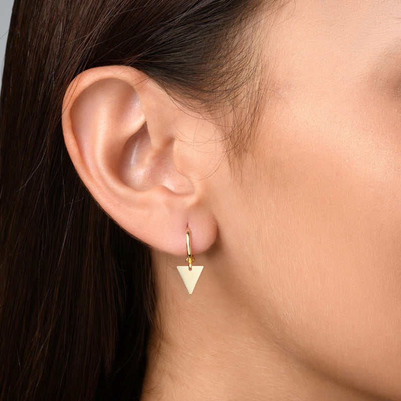 Image of a woman with brown hair wearing gold hoop earrings with a geometric triangle charm. The hoop earrings feature a sleek, minimalist design with a delicate triangle charm that adds a touch of sophistication and style to the woman's look. The gold plating gives the earrings a luxurious and elegant look that complements the woman's brown hair and can be paired with various outfits for a fashionable statement