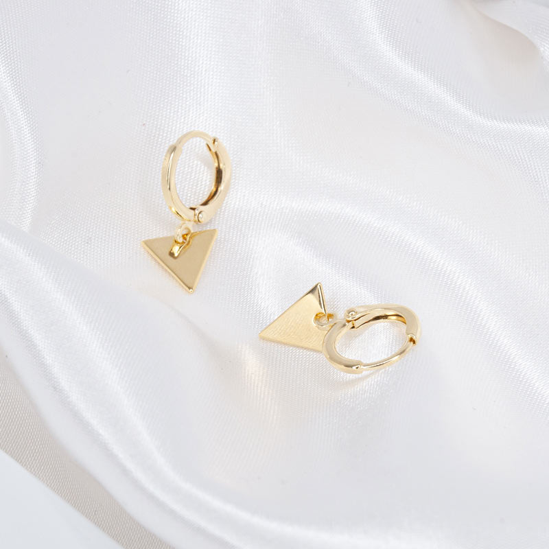 A pair of gold hoop earrings with a triangle charm pendant, lying on a silk fabric. The earrings feature a sleek, minimalist design with a delicate triangle charm that adds a touch of sophistication and style. The image allows viewers to appreciate the fine details and craftsmanship of the earrings, making them an ideal choice for a stylish and fashionable look."