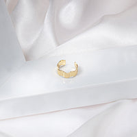 Gold wave-shaped ear cuff designed to fit around the conch of the ear, displayed on a white frame with white silk. The unique, wavy design adds elegance and sophistication. It can be worn alone or combined with other earrings. Clean and modern design