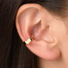 A woman wearing a gold wave-shaped ear cuff designed to fit around the conch of her ear. The ear cuff features a unique, wavy design that adds a touch of elegance and sophistication to the woman's look. It is designed to fit comfortably without requiring a piercing and can be worn on its own or combined with other earrings for a fashionable statement.