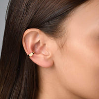 A woman with brown hair wearing a gold wave-shaped ear cuff designed to fit around the conch of her ear. The ear cuff features a unique, wavy design that adds a touch of elegance and sophistication to the woman's look. It is designed to fit comfortably without requiring a piercing and can be worn on its own or combined with other earrings for a fashionable statement.